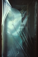 A thin, translucent plastic sheet covers an object beneath, creating a distorted and shadowy silhouette. The surface of the plastic has a frosted appearance with subtle reflections and creases, suggesting texture and depth.