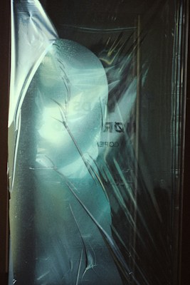 A thin, translucent plastic sheet covers an object beneath, creating a distorted and shadowy silhouette. The surface of the plastic has a frosted appearance with subtle reflections and creases, suggesting texture and depth.