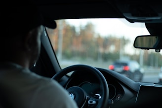 A person is seated in the driver's seat of a BMW vehicle, holding the steering wheel. The view through the windshield shows a blurred background with another car on the road. The interior of the car features a visible side mirror and dashboard.