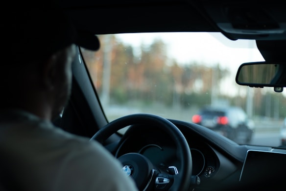 A person is seated in the driver's seat of a BMW vehicle, holding the steering wheel. The view through the windshield shows a blurred background with another car on the road. The interior of the car features a visible side mirror and dashboard.