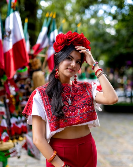 A woman wearing traditional attire with an embroidered red top, matching red pants, and a floral headpiece stands outdoors. Several Mexican flags are in the background, contributing to the festive atmosphere.