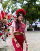 A woman stands outdoors wearing a traditional, colorful outfit with a red floral headpiece. The background includes Mexican flags attached to poles and various items for sale, suggesting a festive or cultural market setting. The woman is adorned with jewelry and her long dark hair is styled in waves.