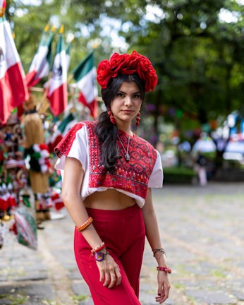 A woman stands outdoors wearing a traditional, colorful outfit with a red floral headpiece. The background includes Mexican flags attached to poles and various items for sale, suggesting a festive or cultural market setting. The woman is adorned with jewelry and her long dark hair is styled in waves.
