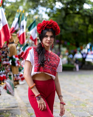 A woman stands outdoors wearing a traditional, colorful outfit with a red floral headpiece. The background includes Mexican flags attached to poles and various items for sale, suggesting a festive or cultural market setting. The woman is adorned with jewelry and her long dark hair is styled in waves.