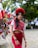 A woman stands outdoors wearing a traditional, colorful outfit with a red floral headpiece. The background includes Mexican flags attached to poles and various items for sale, suggesting a festive or cultural market setting. The woman is adorned with jewelry and her long dark hair is styled in waves.
