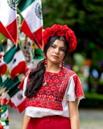 A woman is wearing a traditional embroidered blouse with intricate red and black patterns. She has a red floral headpiece and red earrings, complementing her outfit. Behind her, multiple flags are displayed, showing the colors green, white, and red.