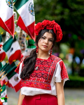 A woman is wearing a traditional embroidered blouse with intricate red and black patterns. She has a red floral headpiece and red earrings, complementing her outfit. Behind her, multiple flags are displayed, showing the colors green, white, and red.