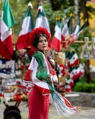 A woman wearing a traditional outfit with a red dress and shawl adorned with green, white, and red colors. She is standing in front of several Mexican flags and colorful decorations in an outdoor setting.