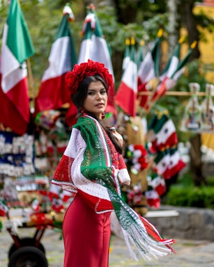 A woman wearing a traditional outfit with a red dress and shawl adorned with green, white, and red colors. She is standing in front of several Mexican flags and colorful decorations in an outdoor setting.
