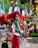 A woman wearing a traditional outfit with a red dress and shawl adorned with green, white, and red colors. She is standing in front of several Mexican flags and colorful decorations in an outdoor setting.
