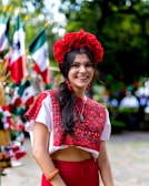 A woman is dressed in traditional clothing, featuring vibrant red and white patterns. She wears a large red floral headpiece and matching earrings. In the background, multiple Mexican flags are visible, suggesting a festive or nationalistic celebration. The setting appears to be outdoors with greenery.