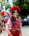 A woman is dressed in traditional clothing, featuring vibrant red and white patterns. She wears a large red floral headpiece and matching earrings. In the background, multiple Mexican flags are visible, suggesting a festive or nationalistic celebration. The setting appears to be outdoors with greenery.