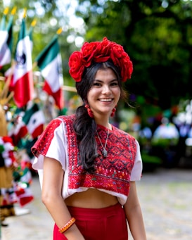 A woman is dressed in traditional clothing, featuring vibrant red and white patterns. She wears a large red floral headpiece and matching earrings. In the background, multiple Mexican flags are visible, suggesting a festive or nationalistic celebration. The setting appears to be outdoors with greenery.