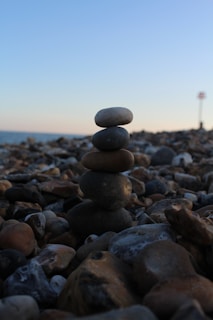 A stack of smooth stones is balanced on a rocky beach, with the ocean and sky in the background. The scene gives an impression of tranquility and balance.