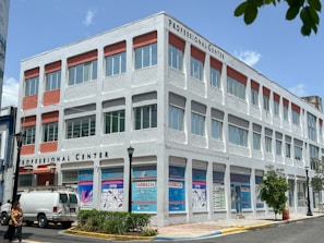 A three-story building labeled 'Professional Center' displays a combination of white and red design elements. The facade features numerous windows and decorative panels. A white van is parked nearby, and signs with text including 'Farmacia' are visible on the ground floor. The scene includes a few people walking, street lamps, and some trees, with a clear blue sky overhead.