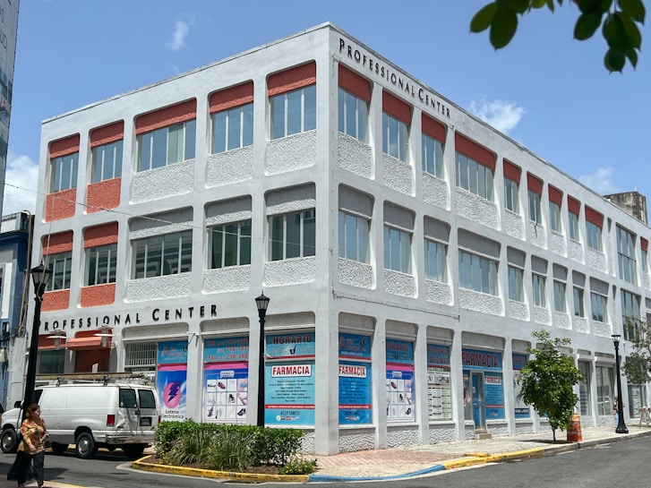 A three-story building labeled 'Professional Center' displays a combination of white and red design elements. The facade features numerous windows and decorative panels. A white van is parked nearby, and signs with text including 'Farmacia' are visible on the ground floor. The scene includes a few people walking, street lamps, and some trees, with a clear blue sky overhead.