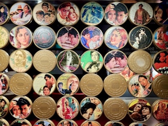A collection of vintage film reel canisters decorated with colorful images from classic movies. The canisters feature a variety of scenes and actors from different films, showcasing vibrant outfits and dramatic expressions.