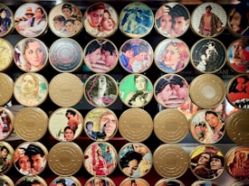 A collection of vintage film reel canisters decorated with colorful images from classic movies. The canisters feature a variety of scenes and actors from different films, showcasing vibrant outfits and dramatic expressions.