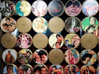 A collection of vintage film reels with covers featuring classic Indian movie posters. The reels are organized in a grid pattern, with colorful and artistic illustrations of actors and scenes from various films. Some reels have labels with titles or quotes.
