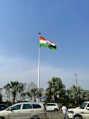 A large flagpole with the Indian flag waving prominently against a clear blue sky. Below, several palm trees and parked cars are visible, along with a person standing near the street.