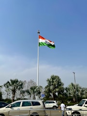 A large flagpole with the Indian flag waving prominently against a clear blue sky. Below, several palm trees and parked cars are visible, along with a person standing near the street.