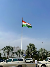 A large flagpole with the Indian flag waving prominently against a clear blue sky. Below, several palm trees and parked cars are visible, along with a person standing near the street.
