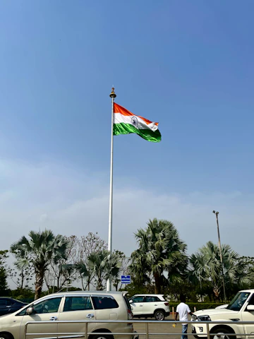 A fleet of buses and cars lined up with the Indian flag waving in the background under a clear sky.