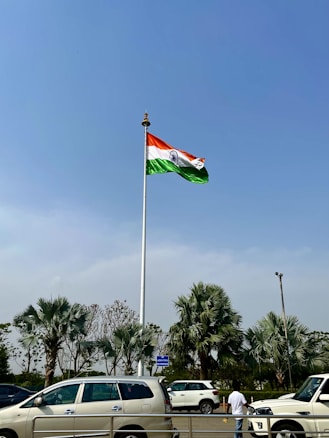 A large flagpole with the Indian flag waving prominently against a clear blue sky. Below, several palm trees and parked cars are visible, along with a person standing near the street.