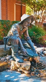 A bronze statue of a man in a hat is kneeling and panning for gold amidst a setting of rocks and flowing water. The setting includes green shrubs, red flowers, and a brick wall backdrop, creating a vibrant outdoor environment.