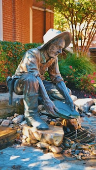 A bronze statue of a man in a hat is kneeling and panning for gold amidst a setting of rocks and flowing water. The setting includes green shrubs, red flowers, and a brick wall backdrop, creating a vibrant outdoor environment.