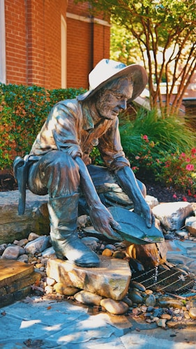 A bronze statue of a man in a hat is kneeling and panning for gold amidst a setting of rocks and flowing water. The setting includes green shrubs, red flowers, and a brick wall backdrop, creating a vibrant outdoor environment.