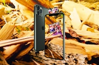 A smartphone is positioned against a backdrop of wooden debris and leaves. The screen displays a colorful card game card. The phone is angled to show both the front screen and the back camera. The surrounding environment includes fragments of wood and autumn leaves.