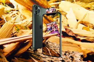 A smartphone is positioned against a backdrop of wooden debris and leaves. The screen displays a colorful card game card. The phone is angled to show both the front screen and the back camera. The surrounding environment includes fragments of wood and autumn leaves.