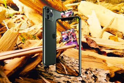 A smartphone is positioned against a backdrop of wooden debris and leaves. The screen displays a colorful card game card. The phone is angled to show both the front screen and the back camera. The surrounding environment includes fragments of wood and autumn leaves.