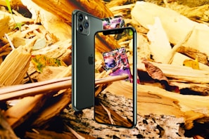 A smartphone is positioned against a backdrop of wooden debris and leaves. The screen displays a colorful card game card. The phone is angled to show both the front screen and the back camera. The surrounding environment includes fragments of wood and autumn leaves.