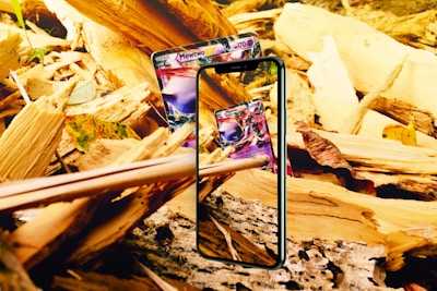 A smartphone is displayed among a bed of broken beige and yellow wood pieces, showcasing a Pokémon card on its screen. Another Pokémon card is situated behind the phone, partially visible. Autumn leaves are scattered among the wooden fragments.