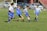 A group of young boys is playing soccer on a grass field. They are wearing blue and white uniforms, and one boy is attempting to kick a soccer ball as others try to intercept. Cars are parked in the background, with a chain-link fence separating the field from the parking area.