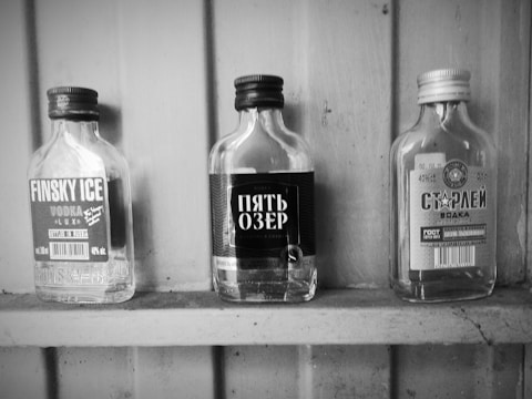 Three empty glass bottles with labels are displayed on a wooden shelf. They include branding in various languages, with prominent text and design elements indicating different vodka brands.