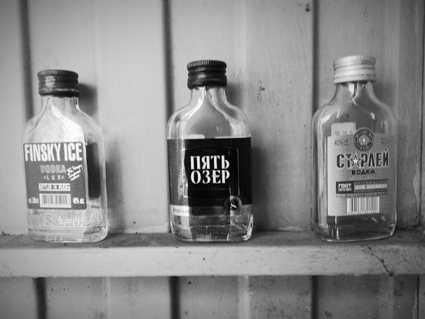 Three empty glass bottles with labels are displayed on a wooden shelf. They include branding in various languages, with prominent text and design elements indicating different vodka brands.