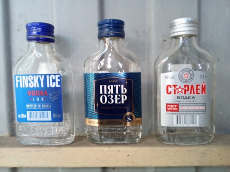 Three small transparent vodka bottles are placed side by side on a shelf against a grey background. The labels on the bottles display different brands and designs. The bottle on the left has a blue label with the text 'Finsky Ice.' The middle bottle has a dark blue label with wavy patterns and Russian text in white. The bottle on the right features a silver label with a red star and additional Russian text.
