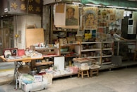A small street market stall featuring a variety of vintage and collectible items. The stall is decorated with numerous posters and artworks, some of which feature historical figures. Shelves are lined with various trinkets, tea sets, framed pictures, and other decorative items. A person stands behind the counter, where more items are displayed.