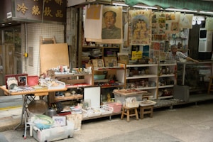A small street market stall featuring a variety of vintage and collectible items. The stall is decorated with numerous posters and artworks, some of which feature historical figures. Shelves are lined with various trinkets, tea sets, framed pictures, and other decorative items. A person stands behind the counter, where more items are displayed.