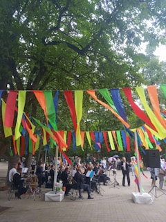 A group of musicians is performing in an outdoor setting, surrounded by trees and festive, colorful streamers hanging overhead. People are gathered around, watching the performance. The scene is lively and vibrant, with a mix of formal attire on the musicians and casual clothing on the spectators.