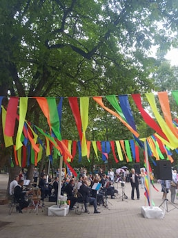 A group of musicians is performing in an outdoor setting, surrounded by trees and festive, colorful streamers hanging overhead. People are gathered around, watching the performance. The scene is lively and vibrant, with a mix of formal attire on the musicians and casual clothing on the spectators.
