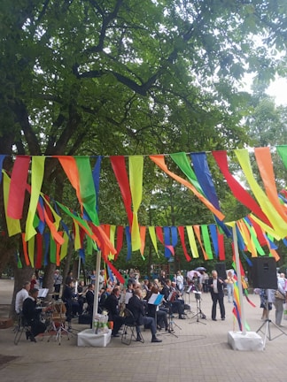 A group of musicians is performing in an outdoor setting, surrounded by trees and festive, colorful streamers hanging overhead. People are gathered around, watching the performance. The scene is lively and vibrant, with a mix of formal attire on the musicians and casual clothing on the spectators.