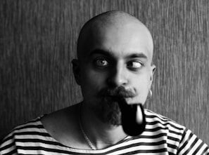 A bald man with a mustache and beard is wearing a striped shirt. He is making a humorous expression by crossing his eyes and has a pipe in his mouth. The background is textured, adding to the contrast in the monochrome image.