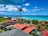 A picturesque coastal view featuring turquoise ocean waters, a clear blue sky with scattered white clouds, and a serene residential area with houses that have red and gray roofs. A tall, singular palm tree stands prominently in the foreground, surrounded by lush green vegetation.