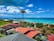 A picturesque coastal view featuring turquoise ocean waters, a clear blue sky with scattered white clouds, and a serene residential area with houses that have red and gray roofs. A tall, singular palm tree stands prominently in the foreground, surrounded by lush green vegetation.