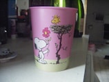 A pink plastic cup with cartoon drawings featuring a dog holding a flower and a bird perched on a nest. The background includes various household items such as bottles and containers.