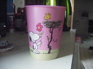 A pink plastic cup with cartoon drawings featuring a dog holding a flower and a bird perched on a nest. The background includes various household items such as bottles and containers.